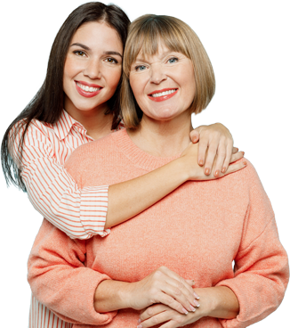 A young woman with long, dark hair smiles brightly while embracing an older woman with short, blonde hair. Both are dressed in casual, cozy outfits, with the older woman wearing a light coral sweater and the younger in a striped, long-sleeved shirt.