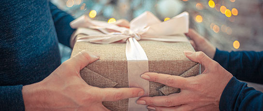 Narrow-Block-Neil Two people holding a gift wrapped with a bow, with twinkling lights in the background.