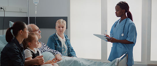 Narrow-Block-drninggao A nurse in blue scrubs speaking with three visitors next to a hospital bed.