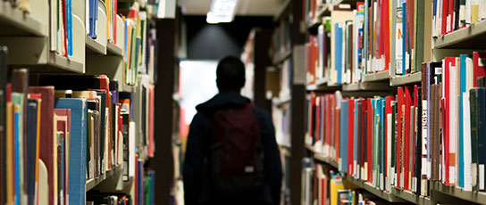 Narrow-Block-laura Person with backpack walking between library bookshelves.