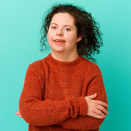 People-Block-26 Person in a rust-coloured sweater with arms crossed against a teal background.