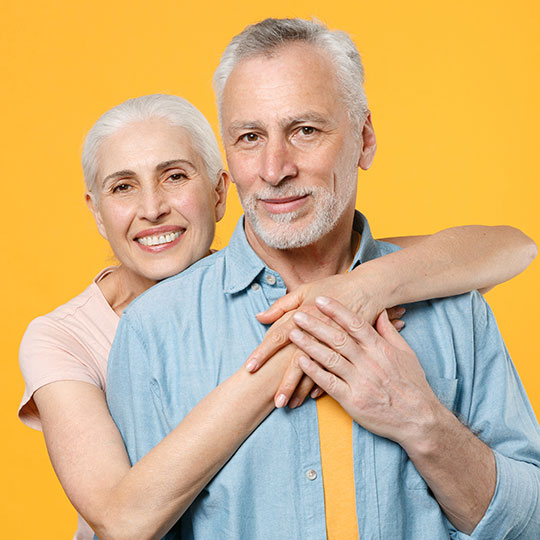 Two senior adults hugging, woman's hands on man's chest, against a yellow background. 