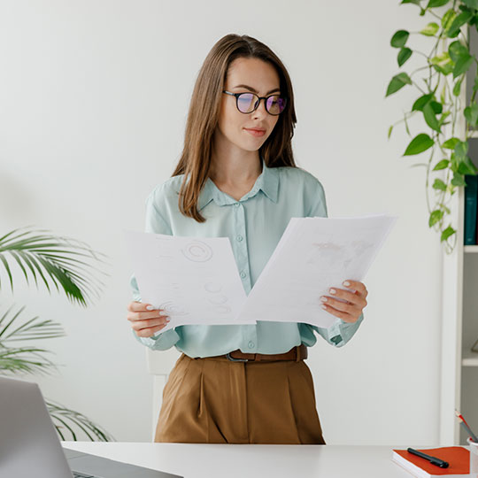 People-Block-29 Woman in a light blue shirt reading documents at a desk with a plant in background.
