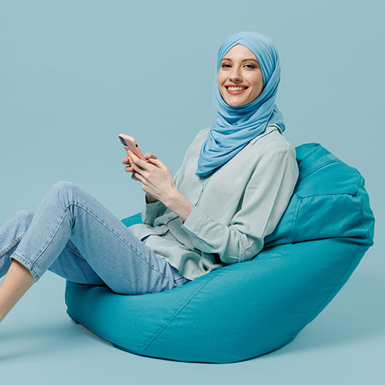 People-Block-8 Person in a blue hijab and outfit sitting on a bean bag chair using a smartphone.